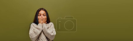 Trendy preteen kid with dyed hair wearing modern cozy knitted sweater while covering face, having fun and standing isolated on green, banner, fashion-forward preteen with sense of style