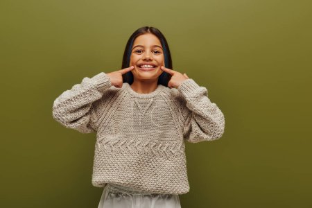 Smiling brunette preteen girl with dyed hair wearing stylish knitted sweater while pointing with finger at mouth and standing isolated on green, fashion-forward preteen with sense of style