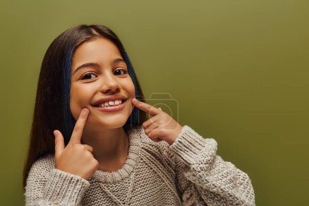 Portrait of cheerful preteen kid with dyed hair wearing modern knitted sweater and pointing with fingers at mouth while standing isolated on green, fashion-forward preteen with sense of style