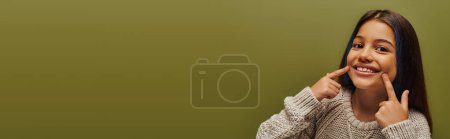 Téléchargez les photos : Enfant préadolescent souriant et à la mode aux cheveux teints portant un pull tricoté tout en pointant les doigts vers la bouche et en se tenant isolé sur un préadolescent vert, bannière, avant-gardiste avec un sens du style - en image libre de droit