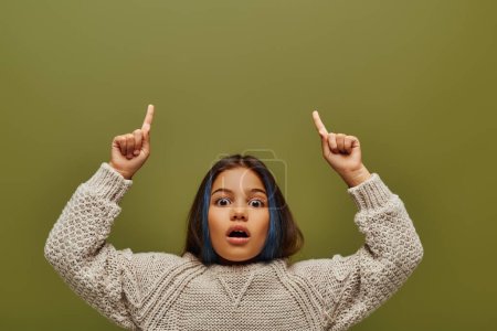 Shocked preteen girl with colored hair wearing stylish knitted sweater while pointing with fingers and looking at camera isolated on green, fashion-forward preteen with sense of style
