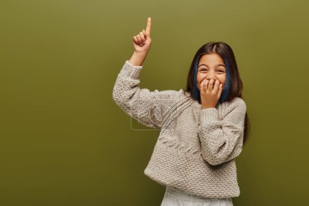 Photo for Laughing and stylish preteen girl with dyed hair wearing knitted sweater and pointing with finger while standing and posing isolated on green, fashion-forward preteen with sense of style - Royalty Free Image