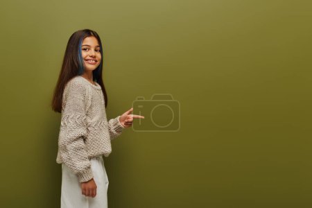 Smiling brunette preteen girl with dyed hair wearing trendy knitted sweater and pointing with finger while looking at camera on green background, fashion-forward preteen with sense of style