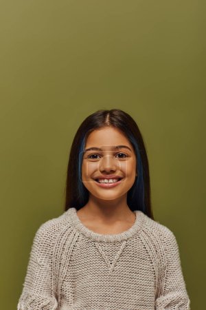 Porträt eines positiven und stilvollen brünetten Mädchens mit gefärbten Haaren, das einen modernen, gemütlichen Strickpullover trägt, während es isoliert auf grünem, modernem Modeschmuck steht und in die Kamera blickt