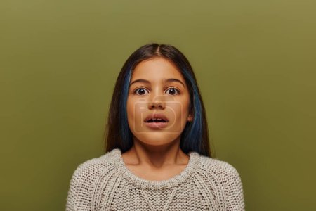 Portrait of scared and stylish preteen girl with dyed hair wearing knitted sweater and looking at camera while standing and posing isolated on green, modern preteen fashion