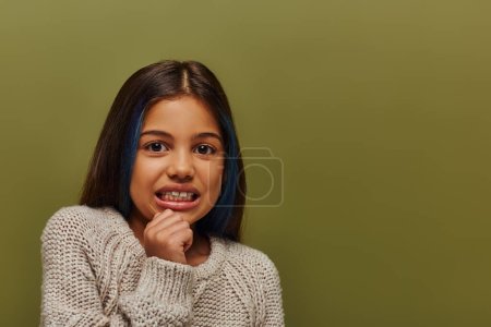 Portrait of disgusted preteen girl with colored hair wearing cozy knitted sweater and looking at camera while standing and posing isolated on green, modern preteen fashion