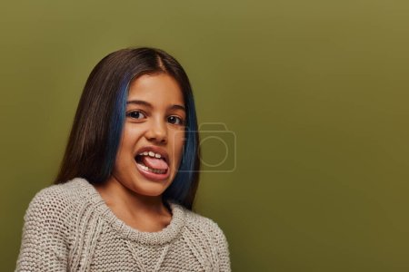 Foto de Retrato de chica preadolescente con estilo con el pelo teñido usando suéter de punto y sobresaliendo lengua mientras se mira a la cámara aislada en verde, moderno y moderno concepto de moda preadolescente - Imagen libre de derechos
