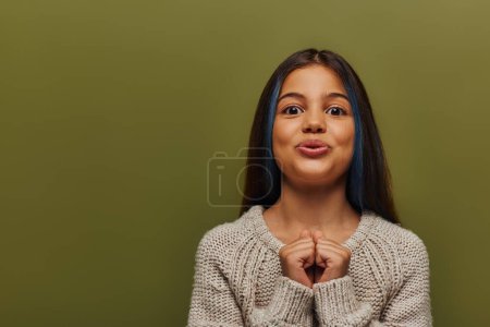 Photo for Excited stylish preteen girl with dyed hair in knitted sweater looking at camera and holding hands near chest while standing and posing isolated on green, stylish girl in cozy fall attire - Royalty Free Image