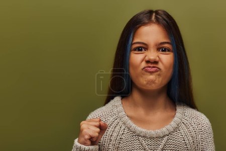 Retrato de niña preadolescente enojada y elegante con el pelo teñido usando suéter de punto mientras mira a la cámara y sosteniendo la mano en el puño aislado en verde, chica elegante en traje de otoño acogedor