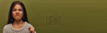 Téléchargez les photos : Portrait d'un préadolescent en colère avec des cheveux teints portant un pull tricoté et regardant la caméra tout en tenant la main dans le poing isolé sur vert, fille élégante en tenue d'automne confortable, bannière avec espace de copie - en image libre de droit