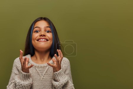 Téléchargez les photos : Fille pré-adolescente excitée avec les cheveux teints portant pull tricoté à la mode tout en gesticulant et en regardant la caméra tout en se tenant isolé sur vert, fille élégante dans le concept de tenue d'automne confortable - en image libre de droit