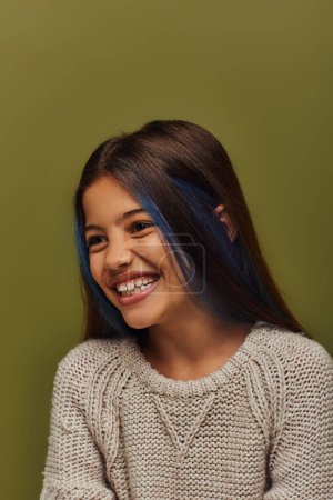 Porträt eines positiven Mädchens mit gefärbten Haaren, das einen modernen und stylischen Strickpullover trägt und wegschaut, während es isoliert auf einem grünen, stylischen Mädchen in gemütlicher Herbstkleidung steht