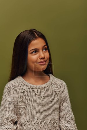 Portrait of pensive and stylish brunette preteen girl in knitted sweater looking away while standing and posing isolated on green, stylish girl in cozy fall attire concept