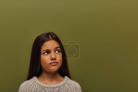 Porträt eines nachdenklichen vorpubertären brünetten Mädchens in stylischem Strickpullover, das im Stehen wegschaut und isoliert auf grünem, stilvollem Mädchen in gemütlichem Herbstkleidungskonzept posiert