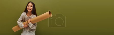 Fröhliches und trendiges Mädchen im modernen Strickpullover mit Papierrollen und Blick in die Kamera, während es isoliert auf grünem, stylisches Mädchen in gemütlicher Herbstkleidung steht, Banner mit Kopierraum