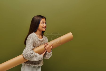 Smiling brunette preadolescent girl in trendy knitted sweater and outfit holding rolled paper and looking away isolated on green, stylish girl in cozy fall attire concept