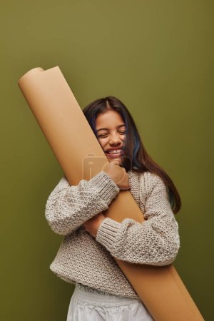 Pleased and stylish preadolescent girl with dyed hair wearing knitted sweater while hugging rolled paper and standing isolated on green, girl radiating autumn vibes concept