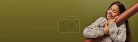Chica preadolescente alegre y elegante con el pelo teñido usando suéter de punto mientras abraza el papel laminado y de pie aislado en verde, niña irradiando vibraciones de otoño, pancarta con espacio para copiar