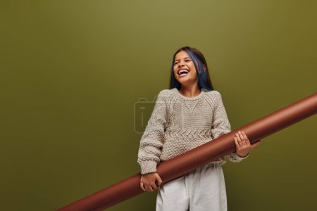 Photo for Excited and stylish preadolescent girl with dyed hair wearing cozy knitted sweater and holding rolled paper while standing isolated on green, girl radiating autumn vibes concept - Royalty Free Image