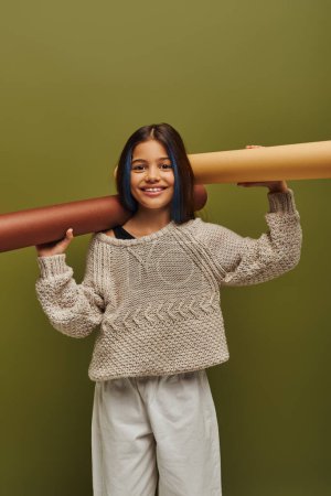 Cheerful preadolescent with dyed hair wearing autumn knitted sweater and looking at camera while holding rolled papers isolated on green, autumn fashion for preteens concept