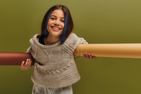 Positive preteen child with dyed hair wearing stylish knitted sweater and autumn outfit looking at camera and holding rolled papers isolated on green, autumn fashion for preteens concept