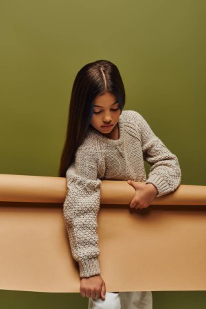 Stylish brunette preadolescent girl in warm and cozy knitted sweater holding rolled paper while standing and posing isolated on green, autumn fashion for preteens concept