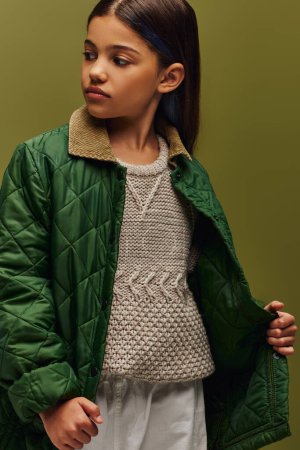 Portrait of trendy brunette preadolescent girl in autumn jacket and modern knitted sweater looking away while standing isolated on green, modern fall fashion for preteens concept