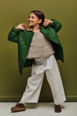 Full length of smiling and trendy preteen kid in autumn outfit and knitted sweater looking away while posing and standing on green background, modern child fashion for preteens concept