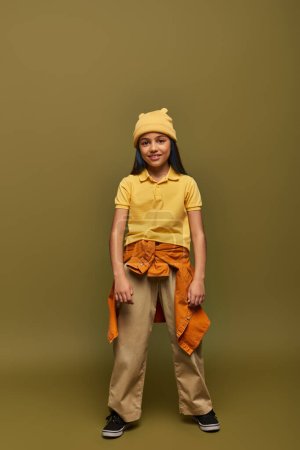Pleine longueur de fille préadolescente souriante et élégante avec des cheveux teints portant une tenue urbaine et un chapeau jaune tout en se tenant debout sur fond kaki, fille élégante dans le concept de tenue moderne
