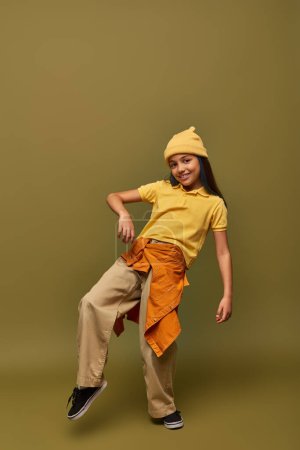 Longitud completa de la chica preadolescente de moda y alegre en traje urbano y sombrero amarillo posando mientras mira a la cámara en el fondo caqui, chica con estilo en el concepto de traje moderno