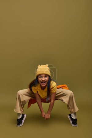 Volle Länge der fröhlichen vorpubertären Mädchen mit gefärbten Haaren tragen gelben Hut und trendige Kleidung posiert, während sie in die Kamera auf khakifarbenem Hintergrund, stilvolles Mädchen in modernem Outfit-Konzept