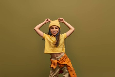 Positive und stilvolle vorpubertäre Mädchen mit gefärbten Haaren tragen urbanes Outfit und berühren gelben Hut, während stehend und posierend isoliert auf Khaki, stilvolles Mädchen in modernem Outfit-Konzept