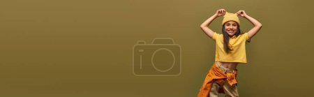 Foto de Sonriente y moderno niño preadolescente con el pelo de color en traje urbano mirando a la cámara mientras toca sombrero amarillo y de pie aislado en color caqui, chica con estilo en el concepto de traje moderno, pancarta - Imagen libre de derechos