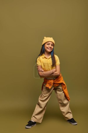Photo for Full length of joyful and stylish preteen girl with dyed hair wearing urban outwear and yellow hat while crossing arms and standing on khaki background, stylish girl in modern outfit concept - Royalty Free Image