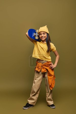 Photo for Full length of cheerful and stylish preteen girl with dyed hair wearing urban outfit and yellow hat and holding skateboard on khaki background, stylish girl in modern outfit concept - Royalty Free Image