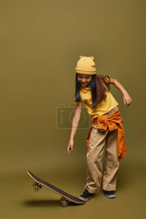 Volle Länge des fröhlichen vorpubertären Kindes mit gefärbten Haaren, das gelben Hut und urbanes Outfit trägt, während es das Skateboard auf khakifarbenem Hintergrund betrachtet, stilvolles Mädchen in modernem Outfit-Konzept