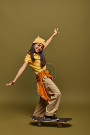 Positive und stilvolle vorpubertäre Mädchen mit gefärbten Haaren tragen gelben Hut und urbanes Outfit, während sie in der Nähe von Skateboard auf khakifarbenem Hintergrund in die Kamera schauen, stilvolles Mädchen in modernem Outfit-Konzept