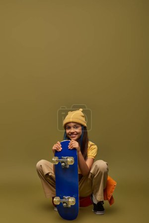 Fashionable preteen girl with dyed hair wearing yellow hat and urban outfit while posing with skateboard and sitting on khaki background, stylish girl in modern outfit concept