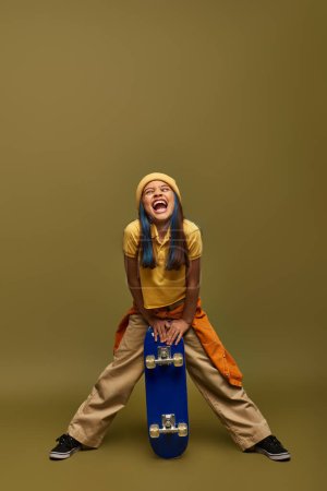 Photo for Full length of excited preteen child with colored hair wearing urban outfit and hat while grimacing and holding skateboard on khaki background, stylish girl in modern outfit concept - Royalty Free Image