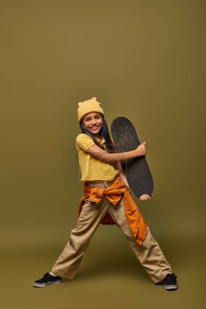 Pleine longueur de fille préadolescente positive en chapeau jaune et tenue urbaine tenant skateboard et regardant la caméra sur fond kaki, fille élégante dans le concept de tenue moderne
