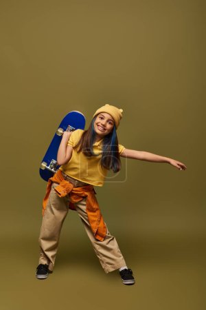 Pleine longueur de fille préadolescente positive avec des cheveux teints portant chapeau jaune et tenue urbaine tout en tenant skateboard et debout sur fond kaki, fille élégante dans le concept de tenue moderne