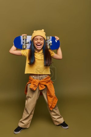Full length of excited preteen kid with dyed hair wearing hat and stylish urban outfit laughing and holding skateboard on khaki background, girl with cool street style look
