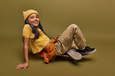 Happy and trendy preadolescent girl with dyed hair wearing urban outfit and yellow hat while looking at camera and sitting on skateboard on khaki background, girl with cool street style look