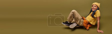 Photo for Positive preadolescent girl with colored hair wearing yellow hat and urban outfit while looking at camera and sitting on skateboard on khaki background, girl with cool street style look, banner - Royalty Free Image