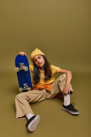 Volle Länge des selbstbewussten und modischen vorpubertären Kindes mit farbigen Haaren, das städtische Kleidung und Hut trägt, während es Skateboard auf khakifarbenem Hintergrund hält, Mädchen mit coolem Streetstyle-Look