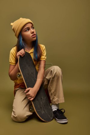 Porträt eines trendigen vorpubertären Mädchens mit farbigen Haaren, das gelben Hut und urbanes Outfit trägt, während es Skateboard hält und auf khakifarbenem Hintergrund wegschaut, Mädchen mit coolem Street-Style-Look