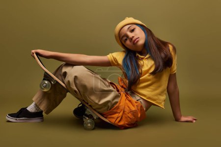 Fashionable preteen girl with dyed hair posing in yellow hat and urban outfit while sitting on skateboard and looking at camera on khaki background, girl in urban streetwear concept