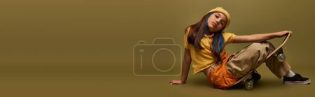 Fashionable preteen kid with dyed hair wearing yellow hat and urban clothes posing and looking at camera while sitting on skateboard on khaki background, girl in urban streetwear concept, banner 