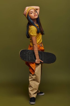 Volle Länge des trendigen und selbstbewussten vorpubertären Kindes mit farbigen Haaren, das Hut und urbanes Outfit trägt und Skateboard hält, während es auf khakifarbenem Hintergrund steht, Mädchen im urbanen Streetwear-Konzept