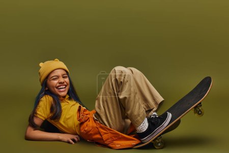 Jeune fille excitée et joyeuse avec des cheveux teints portant des vêtements et un chapeau à la mode tout en étant allongé près du skateboard et en regardant la caméra sur fond kaki, fille dans le concept de streetwear urbain